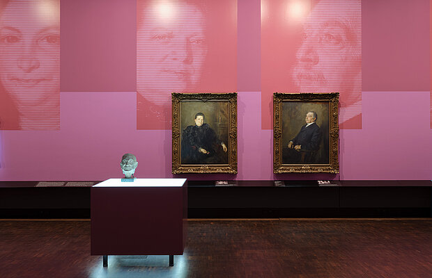 Exhibition view "Picture Stories", portraits of Fanny and Lehmann Bernheimer hanging on a rose-colored wall, in front of them the bust of Otto Bernheimer on an illuminated display case, Photo: Eva Jünger / Jewish Museum Munich