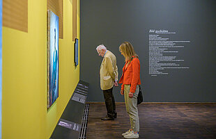 Zwei Besucher*innen in der Ausstellung  „Bildgeschichten“ betrachten zwei an einer hellen gelben Wand hängede Gemälde, Foto: Daniel Schvarcz / Jüdisches Museum München 