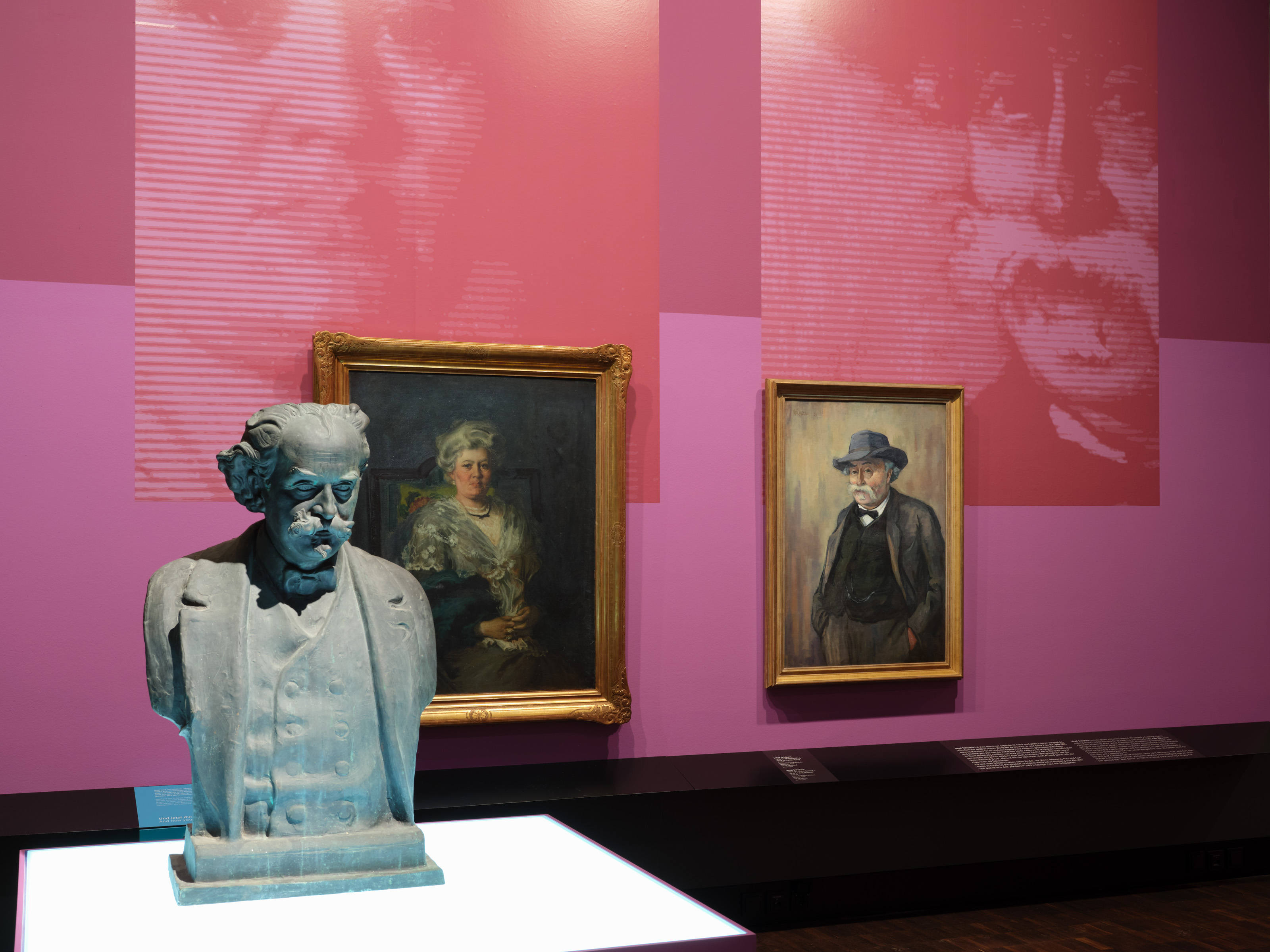 Exhibition view "Picture Stories. Portraits of Munich Jews" - a bust in the foreground and two portraits in the background hang side by side in an exhibition room in shades of pink, large blow-ups reflect the motif of the portraits, photo: Eva Jünger / Jewish Museum Munich
