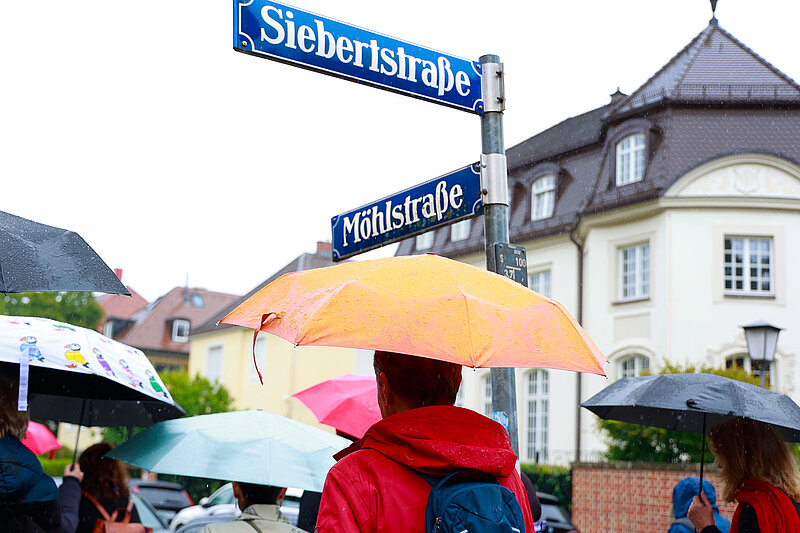 Rundgang des JMM an der Sieberstraße Ecke Möhlstraße Rundgangsteilnehmer*innen mit Regenschirmen stehen unter den Straßenschildern Siebertstraße Ecke Möhlstraße